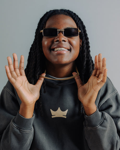 Milaysia Fulwiley wearing sunglasses with a thick chain and a dark Unrivaled hoodie with a crown logo on a gray background