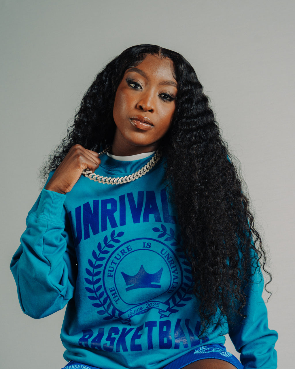 Flau'jae Johnson wearing a blue sweatshirt with 'Unrivaled Basketball', sitting on a chair against a plain background.