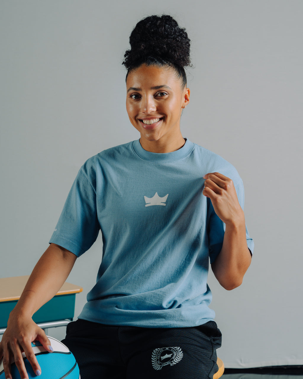 Juju Watkins wearing a blue Unrivaled t-shirt with a crown logo, sitting on a chair against a gray background