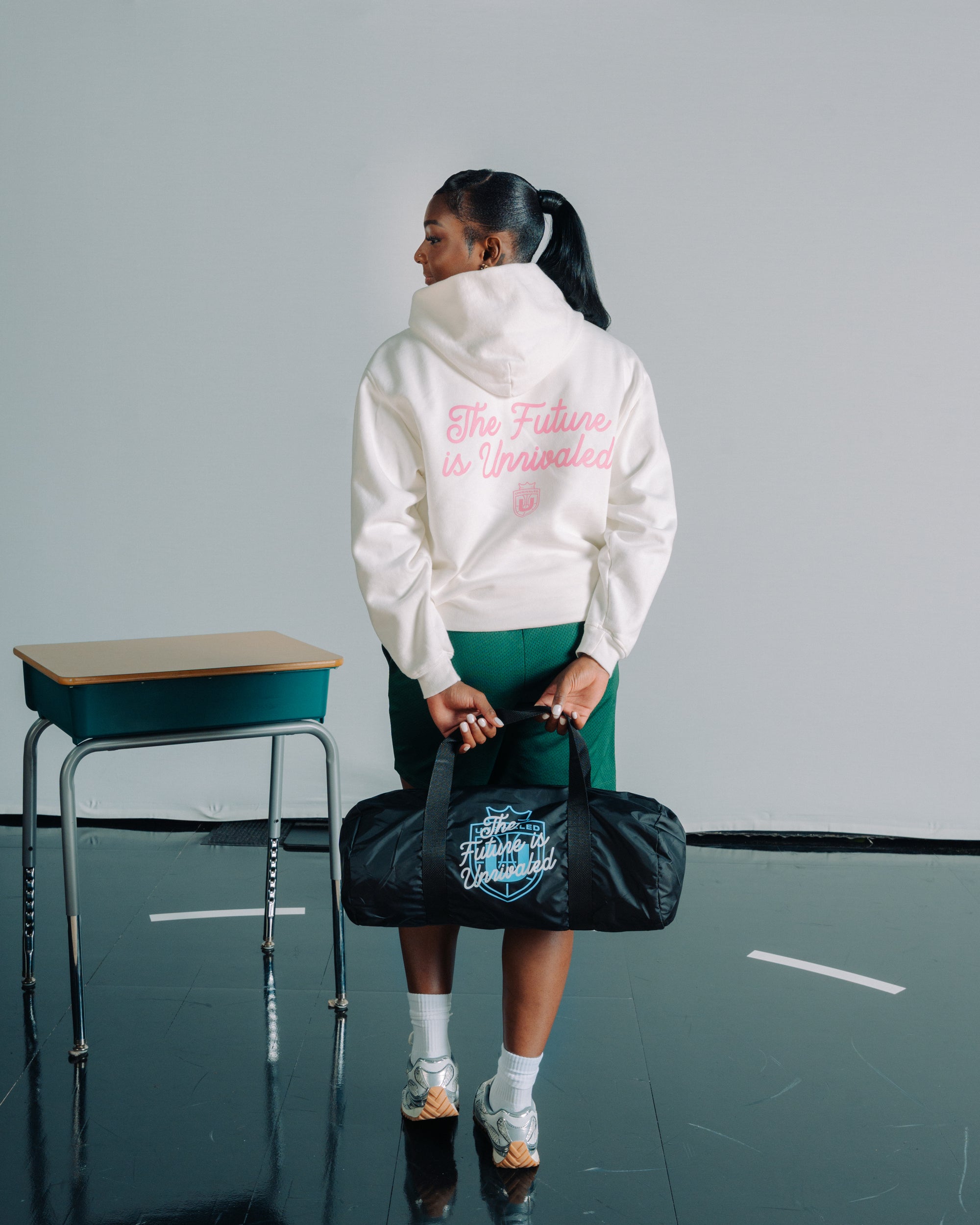 Ta’Niya Latson wearing a white hoodie with text, holding a black duffel bag in an indoor setting.
