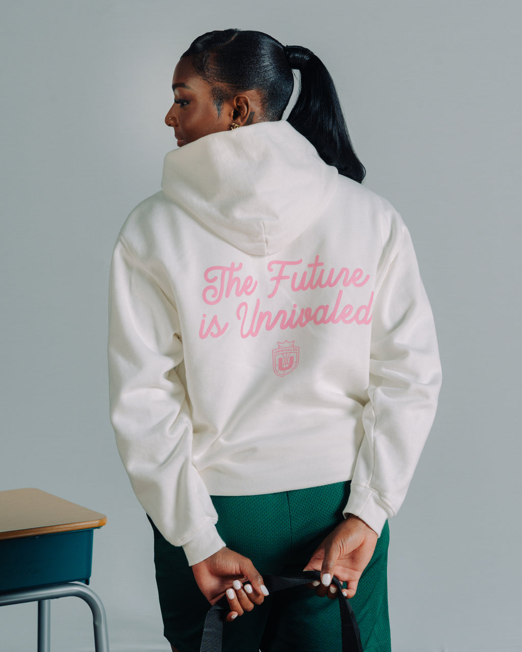 Ta’Niya Latson wearing a white hoodie with 'The Future is Unrivaled' text on the back, standing against a gray background.