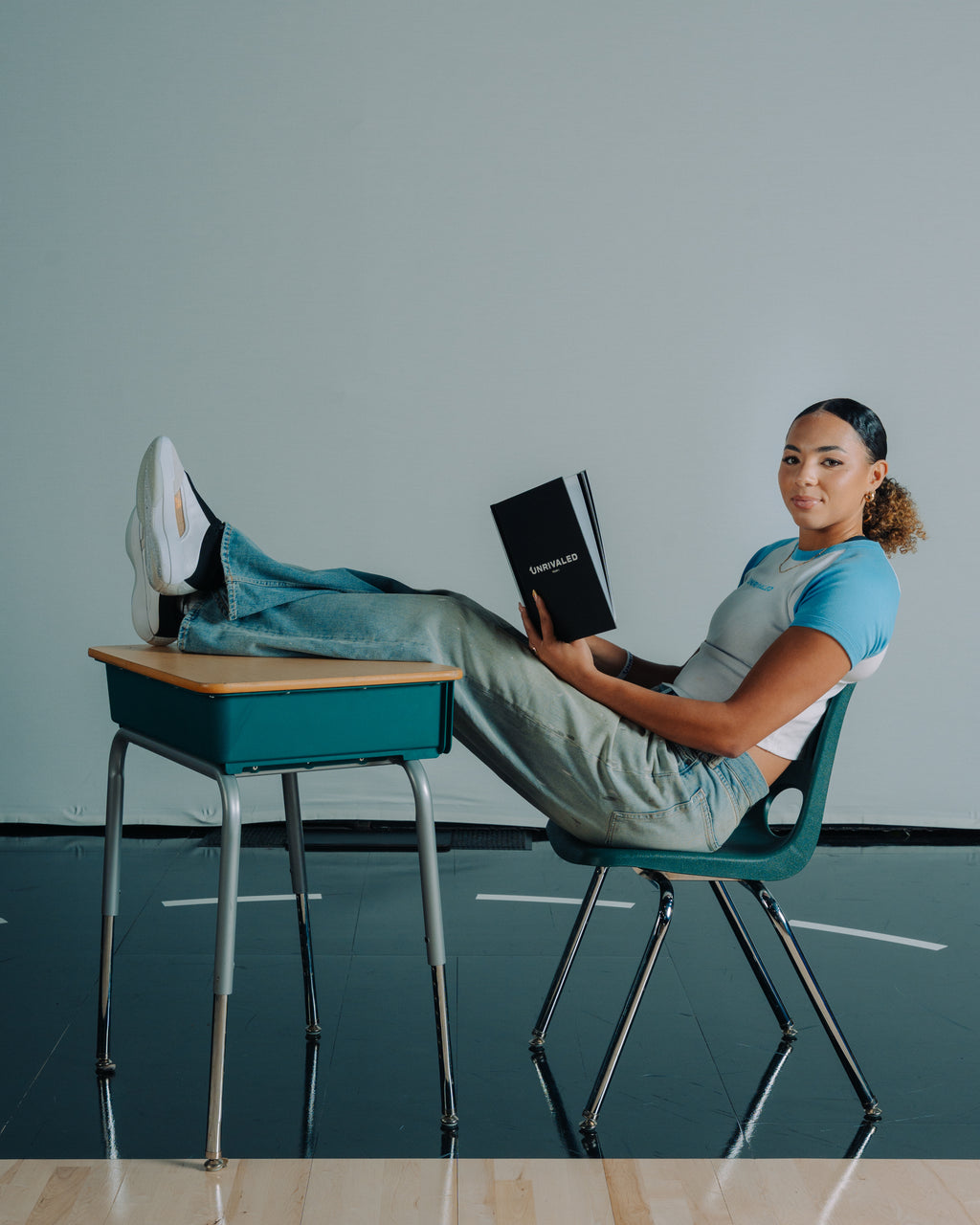 Kiki Rice sitting on a chair with feet on a desk, holding a book in a baby blue unrivaled Tee