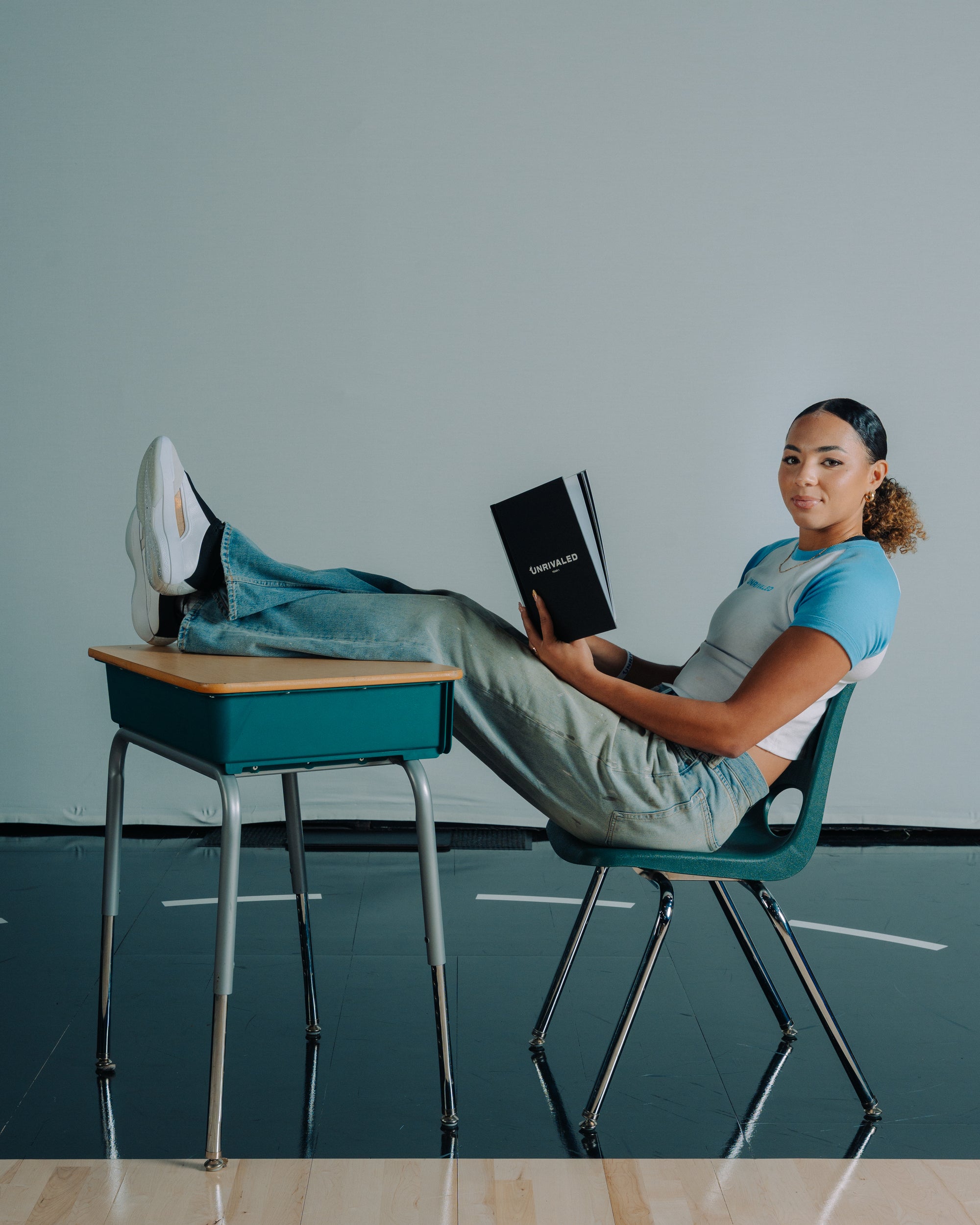 Kiki Rice sitting on a chair with feet on a desk, holding a book in a baby blue unrivaled Tee